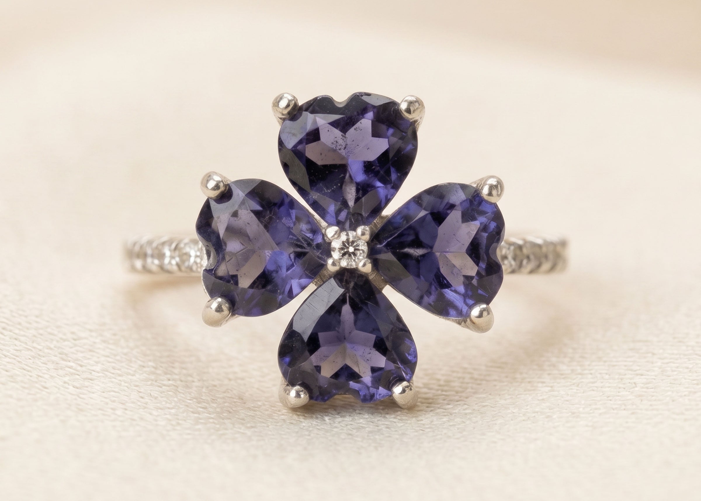 A silver ring featuring a four-petal flower design with heart-shaped deep purple gemstones and a small clear crystal in the center, set on a light cream background.